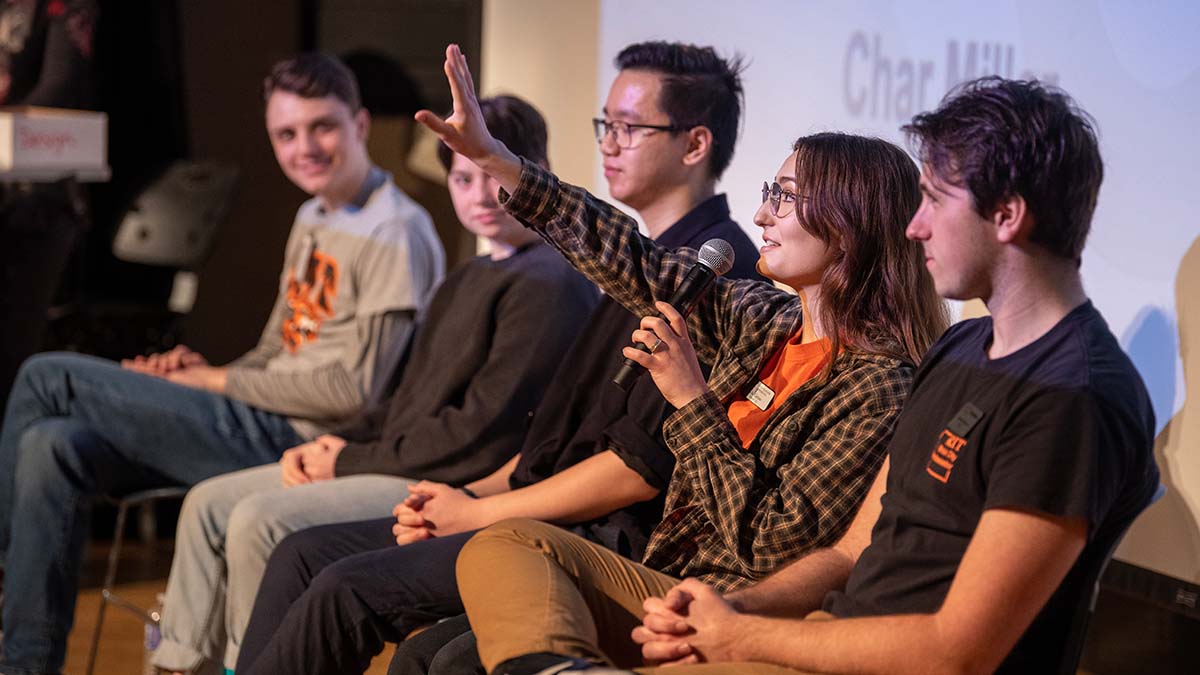 Panelists seated in a row, with one participant raising their hand while speaking into a microphone.