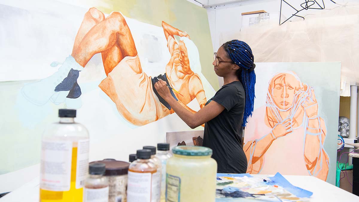 An art student painting a large portrait of a reclining female figure on canvas, surrounded by bottles of paint and materials.