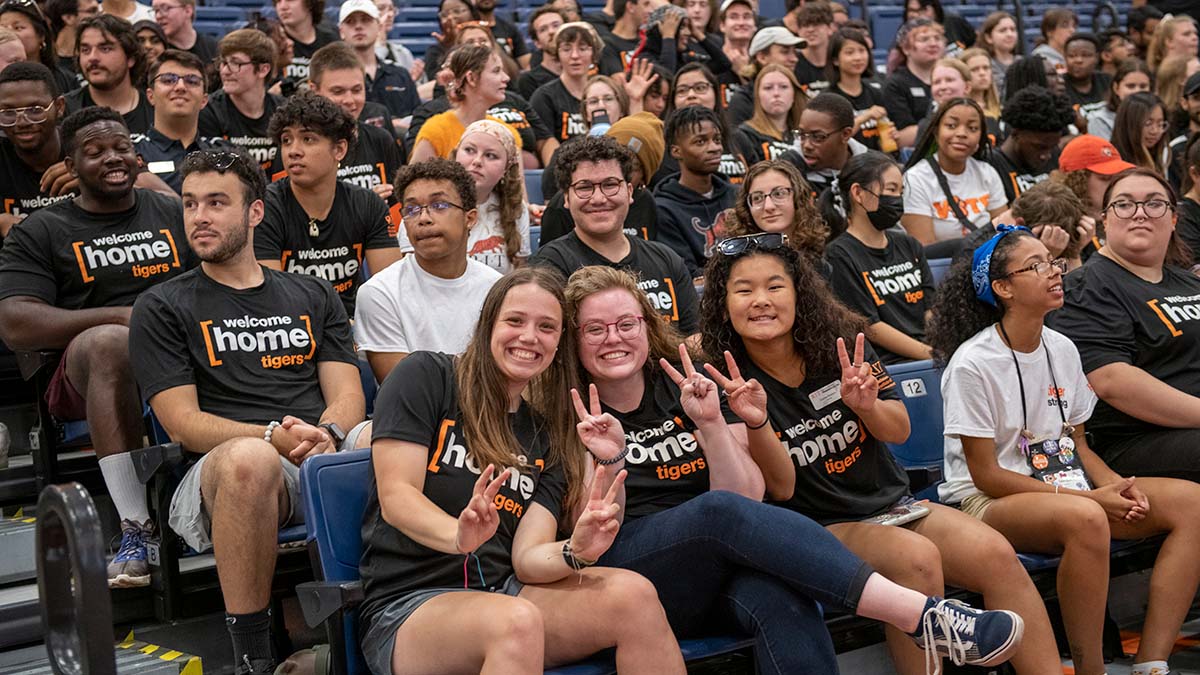 A large group of students wearing 'welcome home tigers' shirts, smiling and seated together at an indoor event.