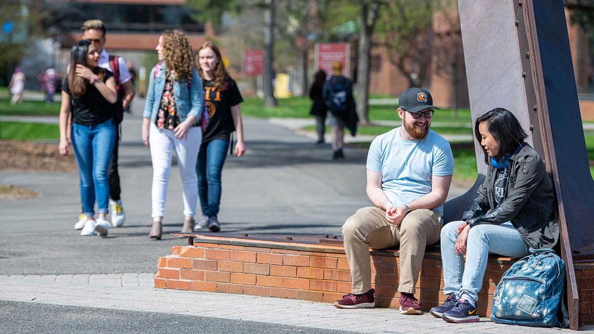 Two students sitting on a low brick wall talking, while others walk along a campus path in the background.