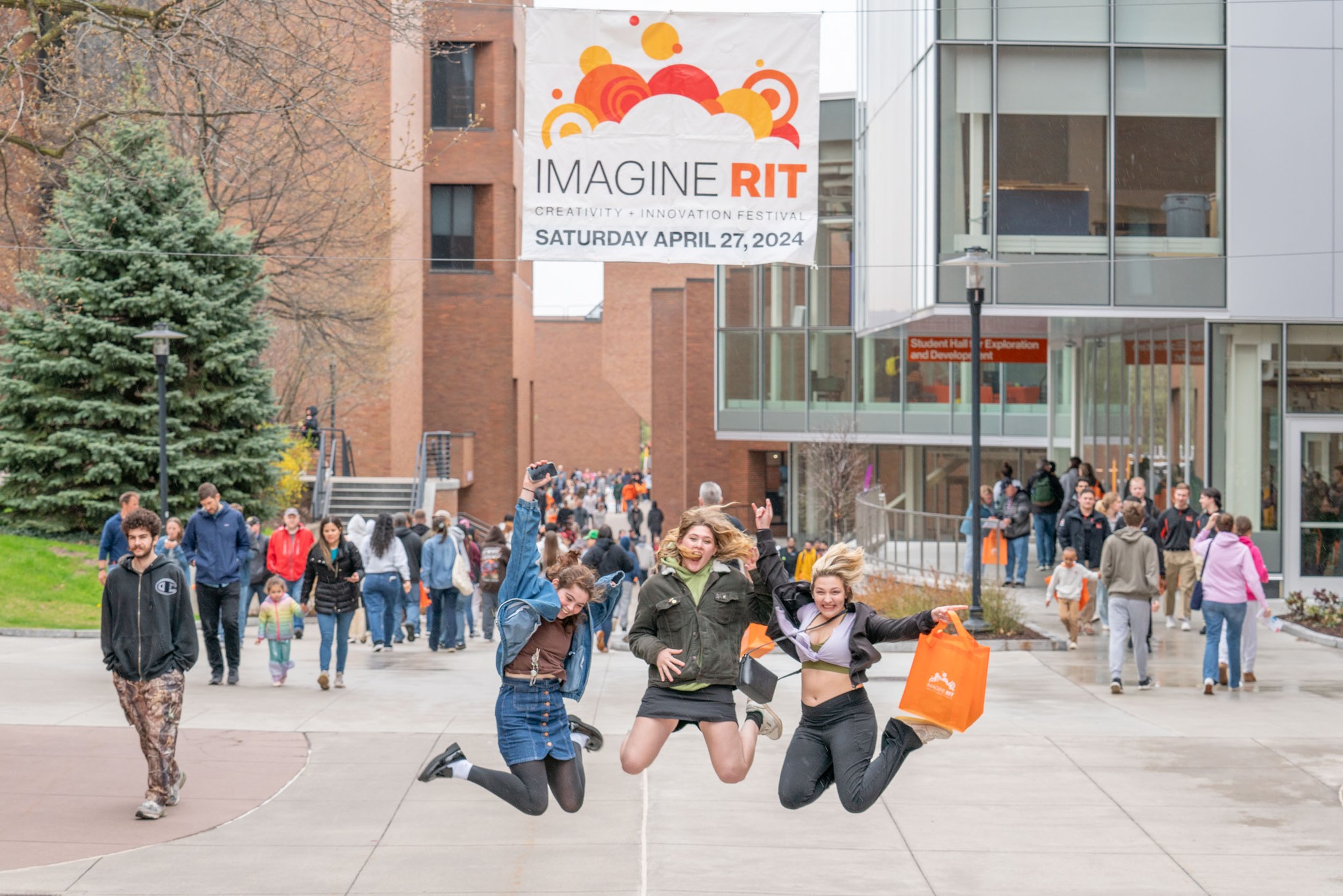 three students jumping up underneath an Imagine RIT banner suspended above a walking path