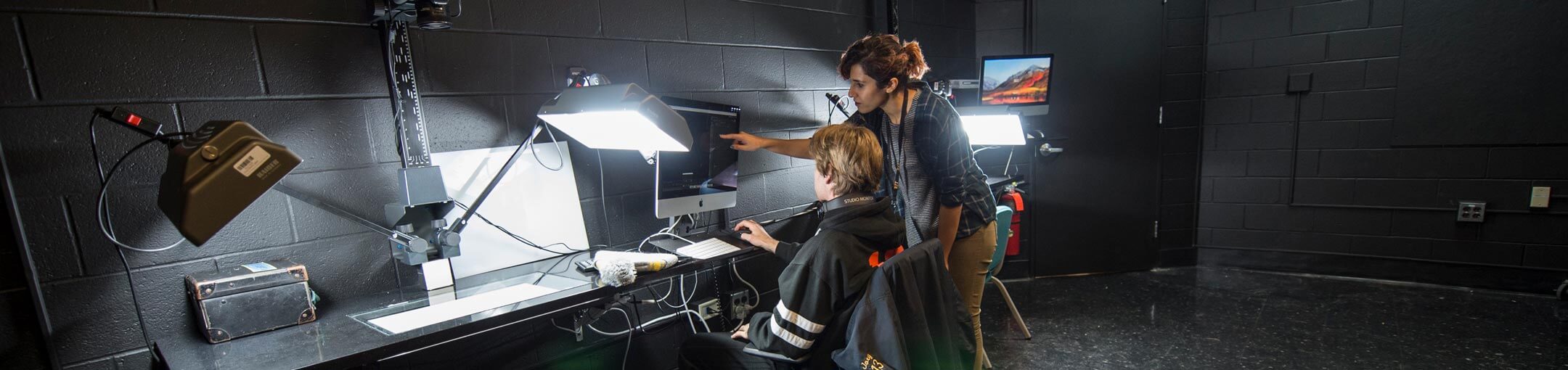 two students looking at a computer screen in a dark studio environment.