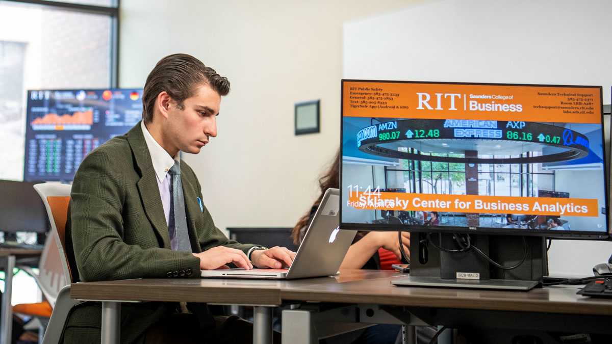 A student works at a laptop with a nearby computer monitor displaying RIT Saunders College of Business on it.