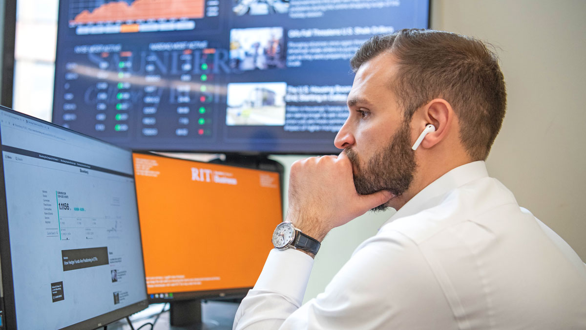 A bearded student wearing ear buds works at a computer.