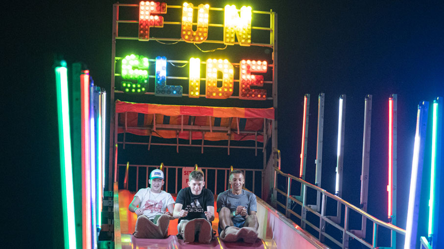 Three R I T students sliding down a carnival slide at night, flanked by neon lights.