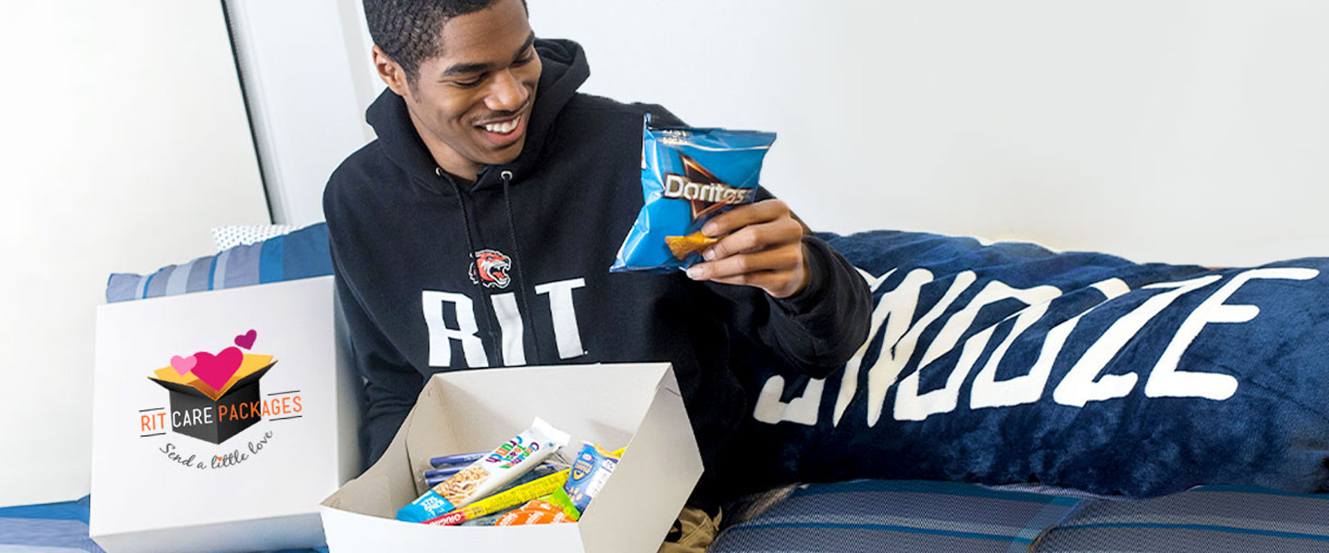 A person sitting on a bed with a black hoodie featuring an RIT logo, holding a blue bag of Doritos, with an open care package box labeled “RIT CARE PACKAGES” filled with various snacks.