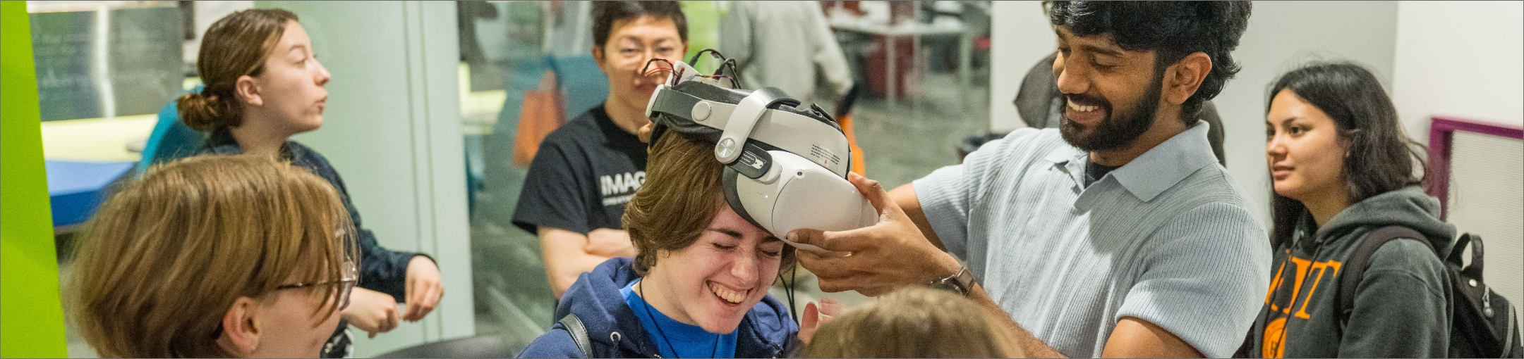 a group of people standing around two individuals one placing a VR headset on the other.