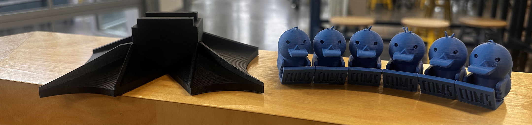 A black 3D-printed object is placed next to a row of blue 3D-printed duck figurines on a wooden surface in a workshop setting.