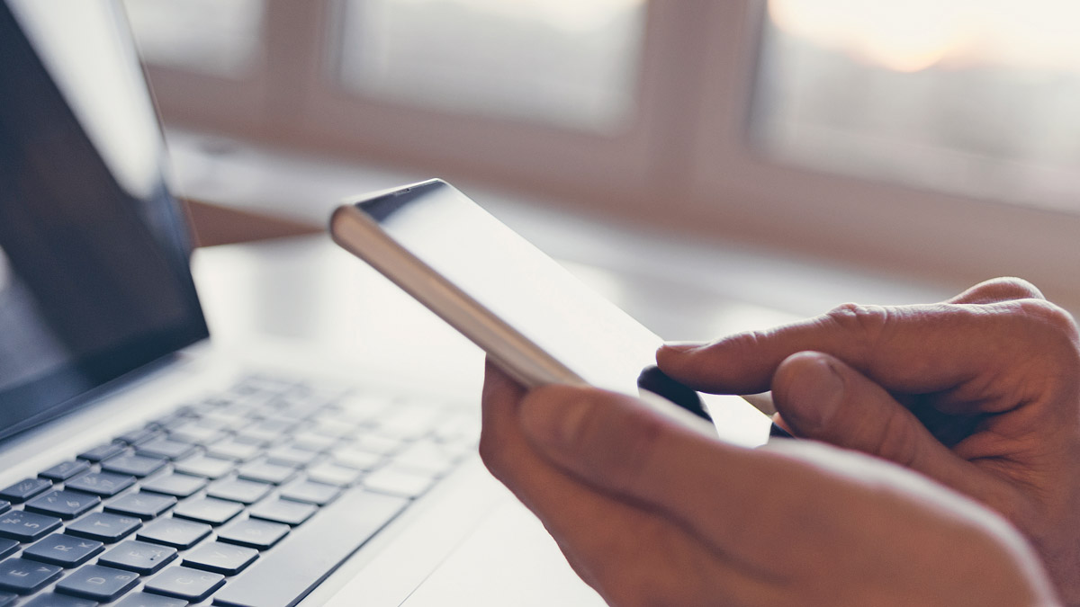 Close up of a finger touching a mobile phone with a laptop out of focus in the background.