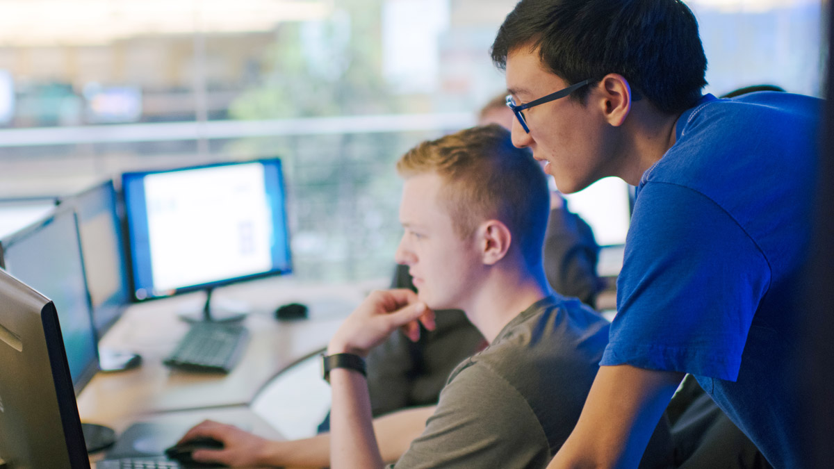 A sitting student and standing student look at computer monitors.