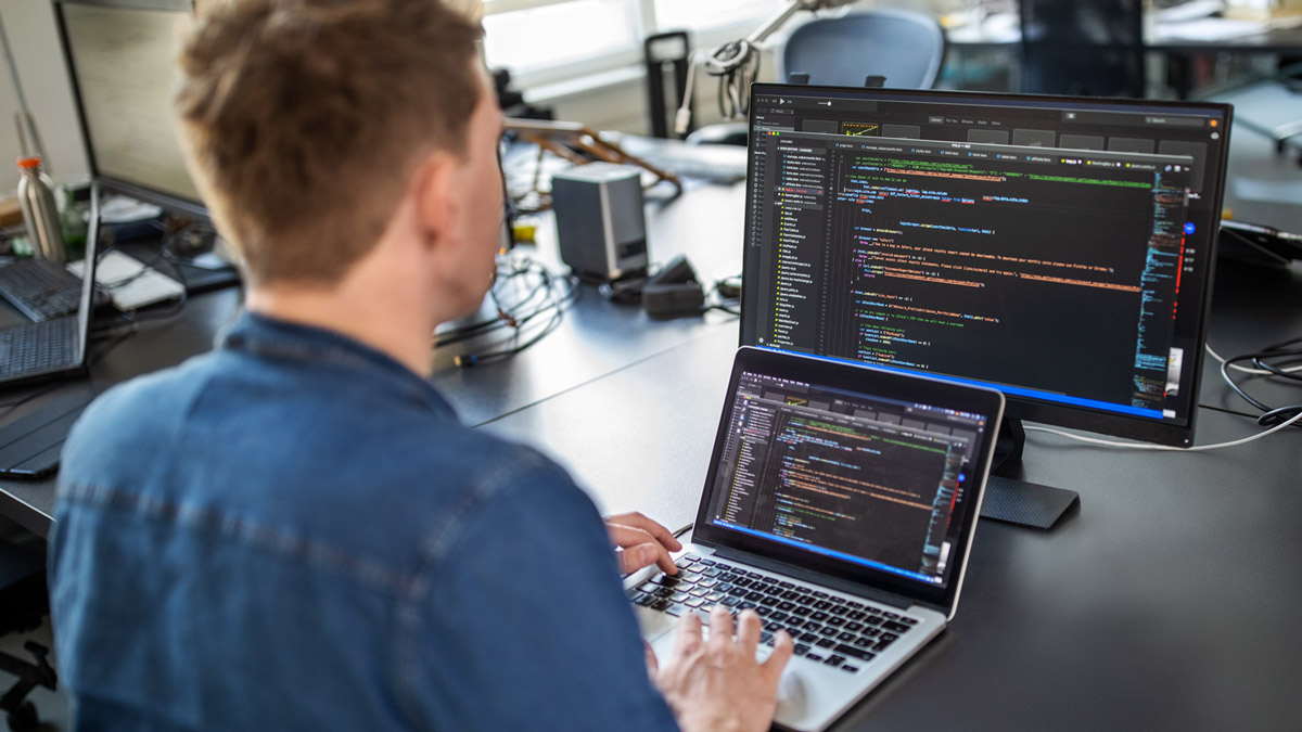 Student sits at a laptop with computer code on it. A monitor also displaying code is behind the laptop.