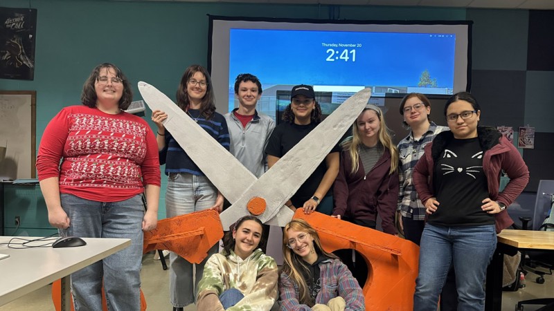 A group of 9 students surrounds a pair of comically oversized scissors constructed from fabric and cardboard
