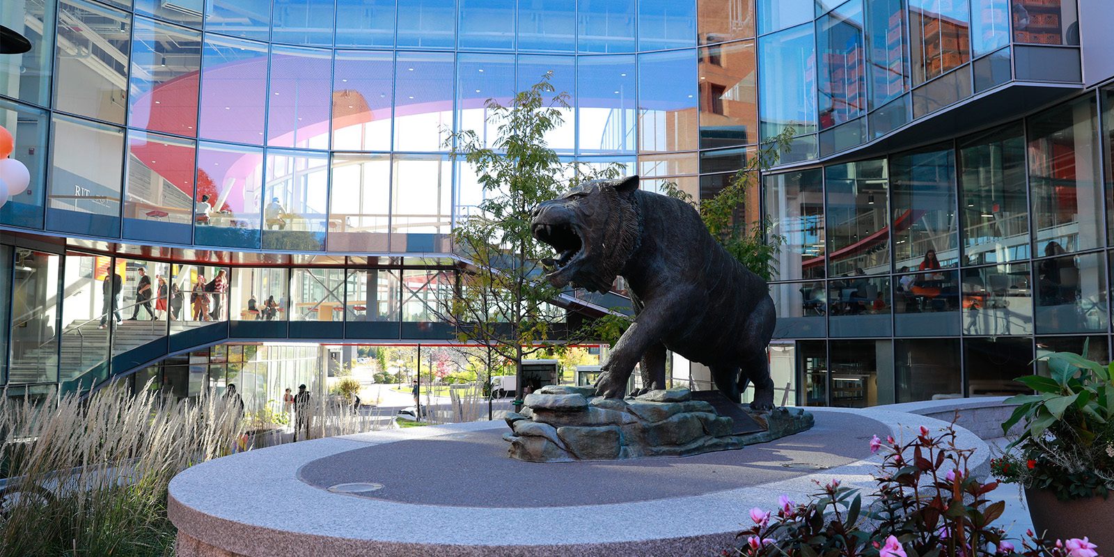 ﻿RIT tiger statue in a modern glass atrium surrounded by plants and reflections of people inside the building