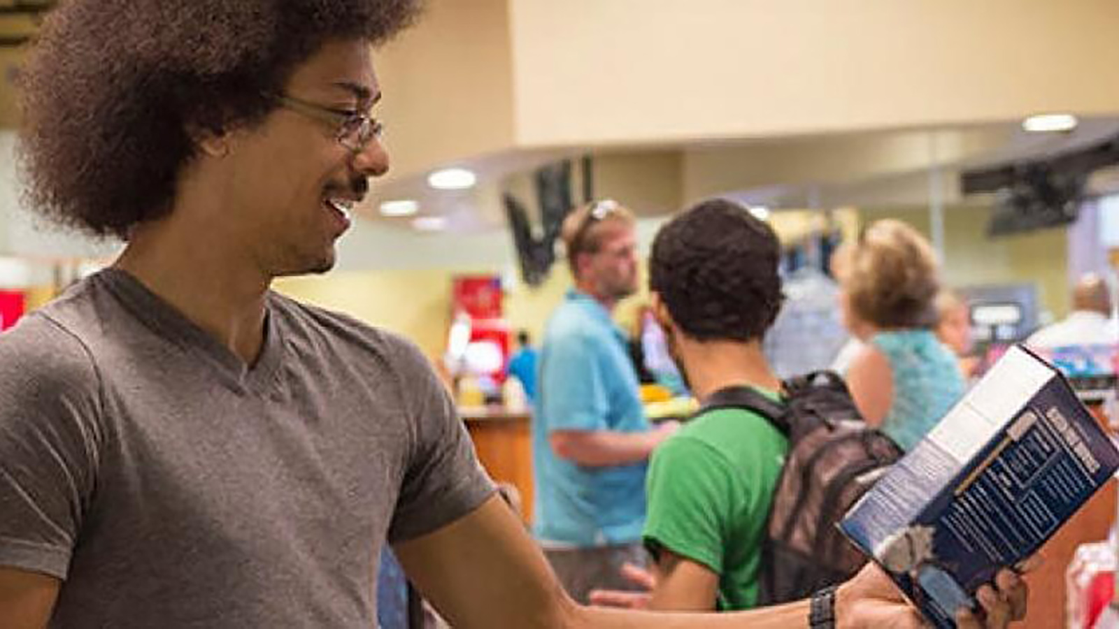 A group of individuals in a public setting, possibly a store or service counter. One person in the foreground is holding an object that looks like a book or pamphlet.