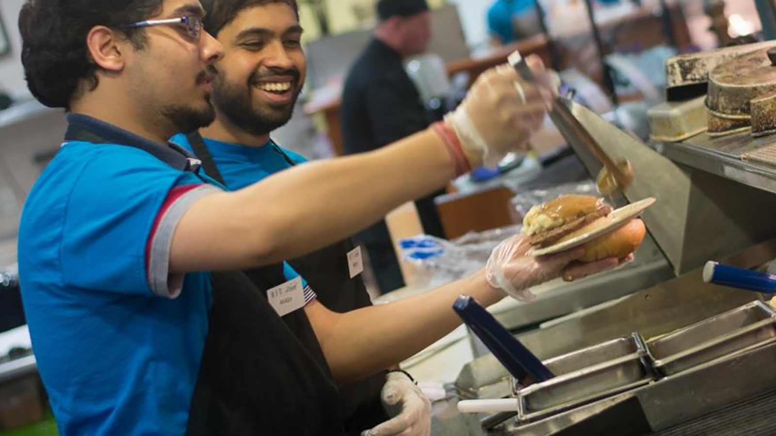 CR--2_CROPPED.txt  An individual in a blue shirt and gloves, standing behind a counter, preparing food. The person is holding a plate with a burger and adding sauce from a spoon. Condiment containers and other food preparation items are on the counter.