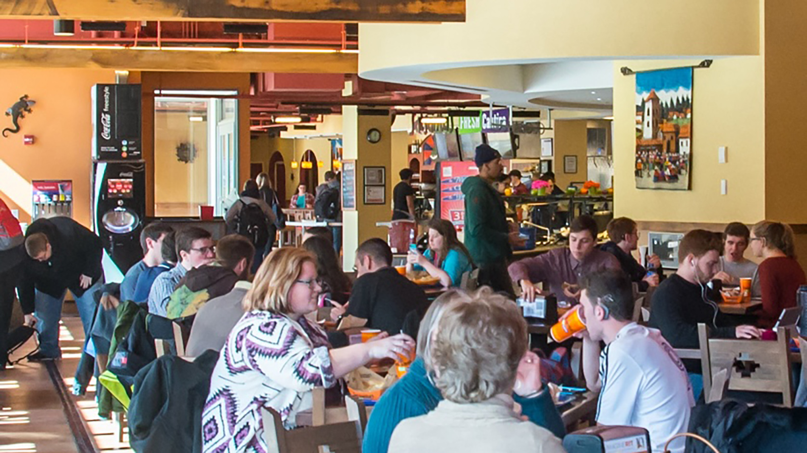 A busy cafeteria or dining area with multiple tables where individuals and groups are seated, eating, and conversing. The room has a high ceiling with exposed beams, a counter at the back for ordering food, and artwork on the wall. Natural light is coming in from windows not visible in the frame.