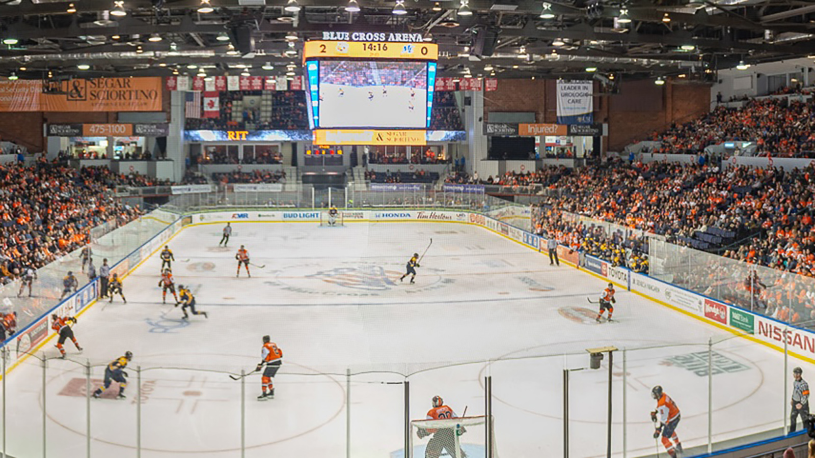 Hero_2160x510_Concessions_0_CROPPED.txt An indoor ice hockey arena during a game, with players on the ice mid-play. The rink is surrounded by boards and protective glass, and the arena is filled with spectators. A scoreboard shows a score of 2-0 with 14:16 remaining in the period.