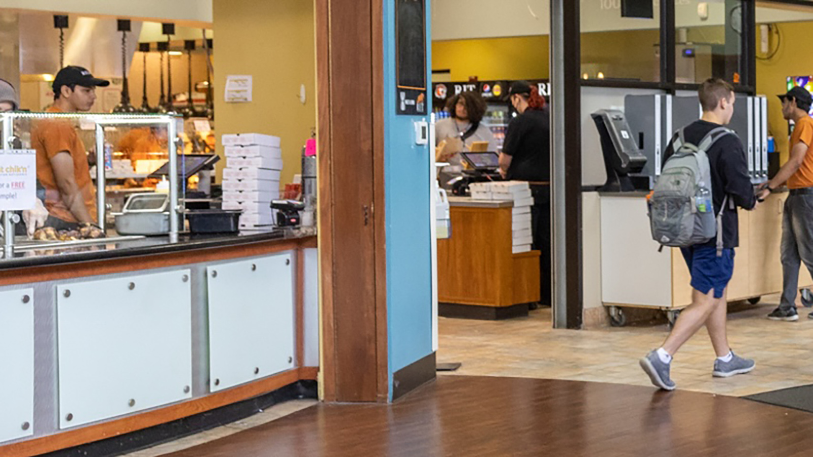 An indoor cafeteria or food court with counters displaying food. One person wearing a cap is standing behind the counter, possibly preparing or serving food. Another individual, wearing a backpack and shorts, is facing away from the camera, selecting their meal.