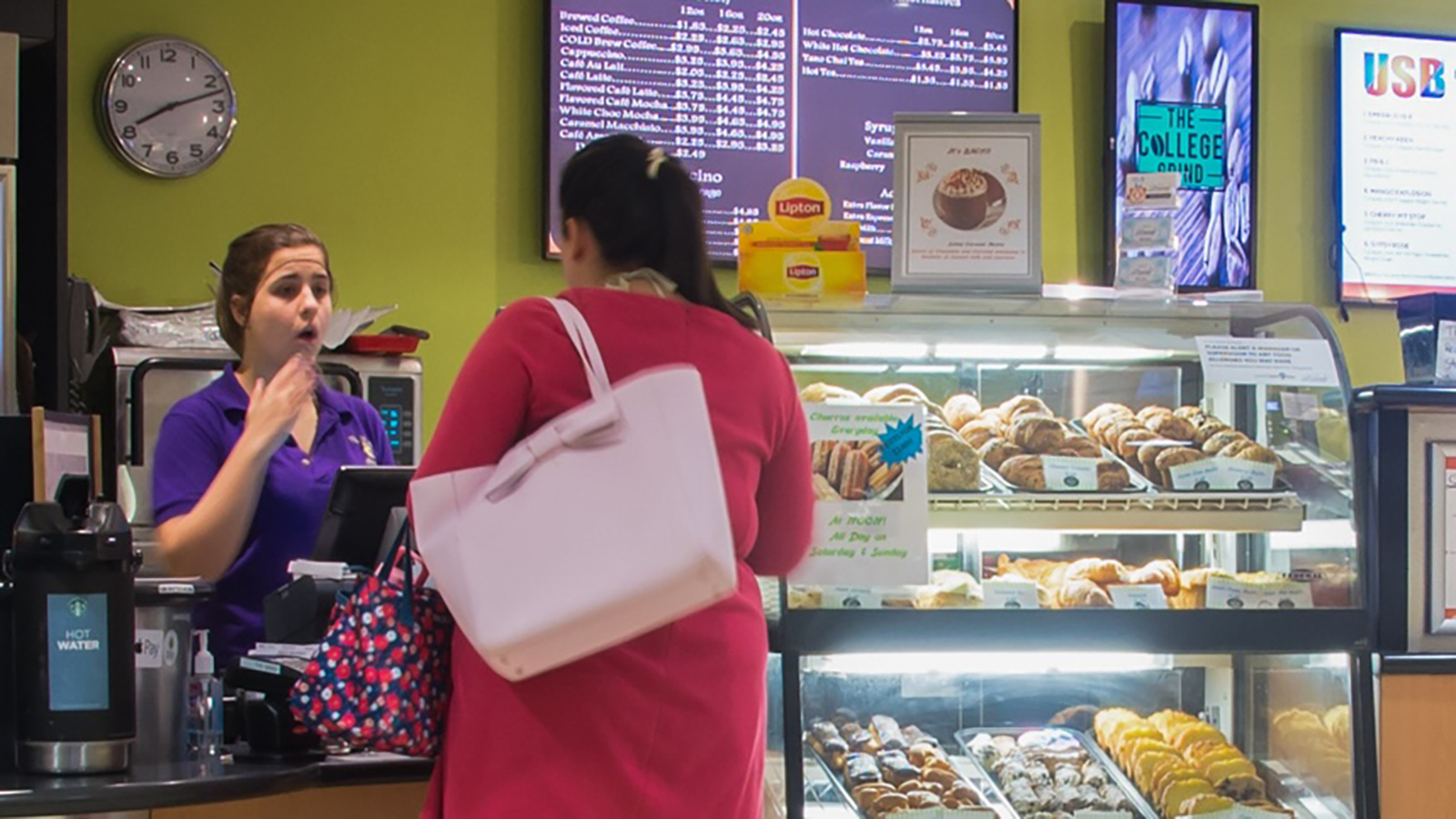 An indoor food establishment with a customer standing at the counter facing an employee. Behind the counter, there is a display of various baked goods and a menu board listing different items and prices. A wall clock shows the time as 10:10, and there are posters or digital screens displaying advertisements or information above the menu board.