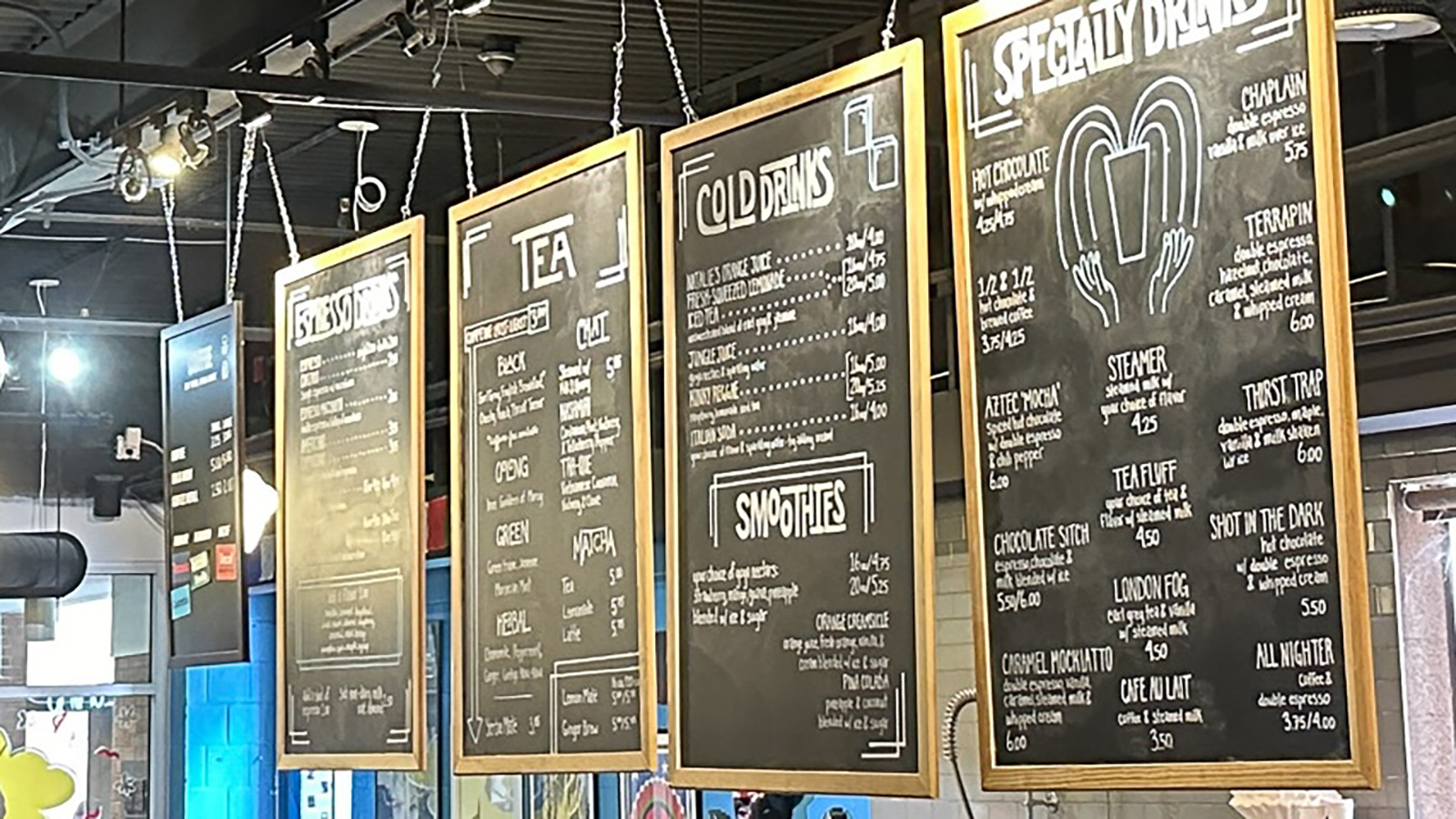 A series of four vertical menus hanging from the ceiling in a cafe or restaurant, listing categories such as “SANDWICHES,” “TEA,” “COLD DRINKS,” and “SMOOTHIES.” The items are written in white on a black background.