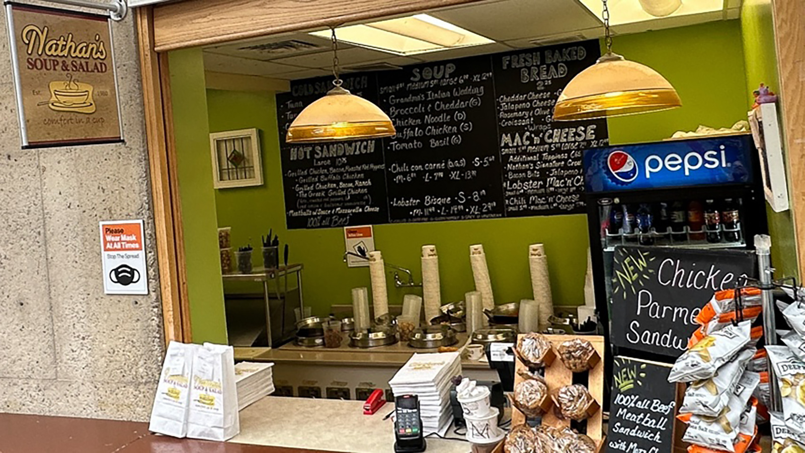 The interior of a food establishment with a counter where orders are taken and prepared. Above the counter, a menu board lists various food items such as soups, salads, hot sandwiches, and mac & cheese. Beverage options like Pepsi are also visible. The area is illuminated by hanging lamps, and various food packaging materials are on the counter.