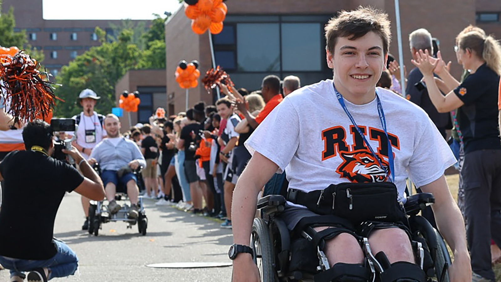 A group of individuals participating in an outdoor event, with some using wheelchairs. One person in the foreground is wearing a t-shirt with the “RIT” logo. The background features other participants and spectators, some holding orange pompoms, suggesting a celebratory or supportive atmosphere.