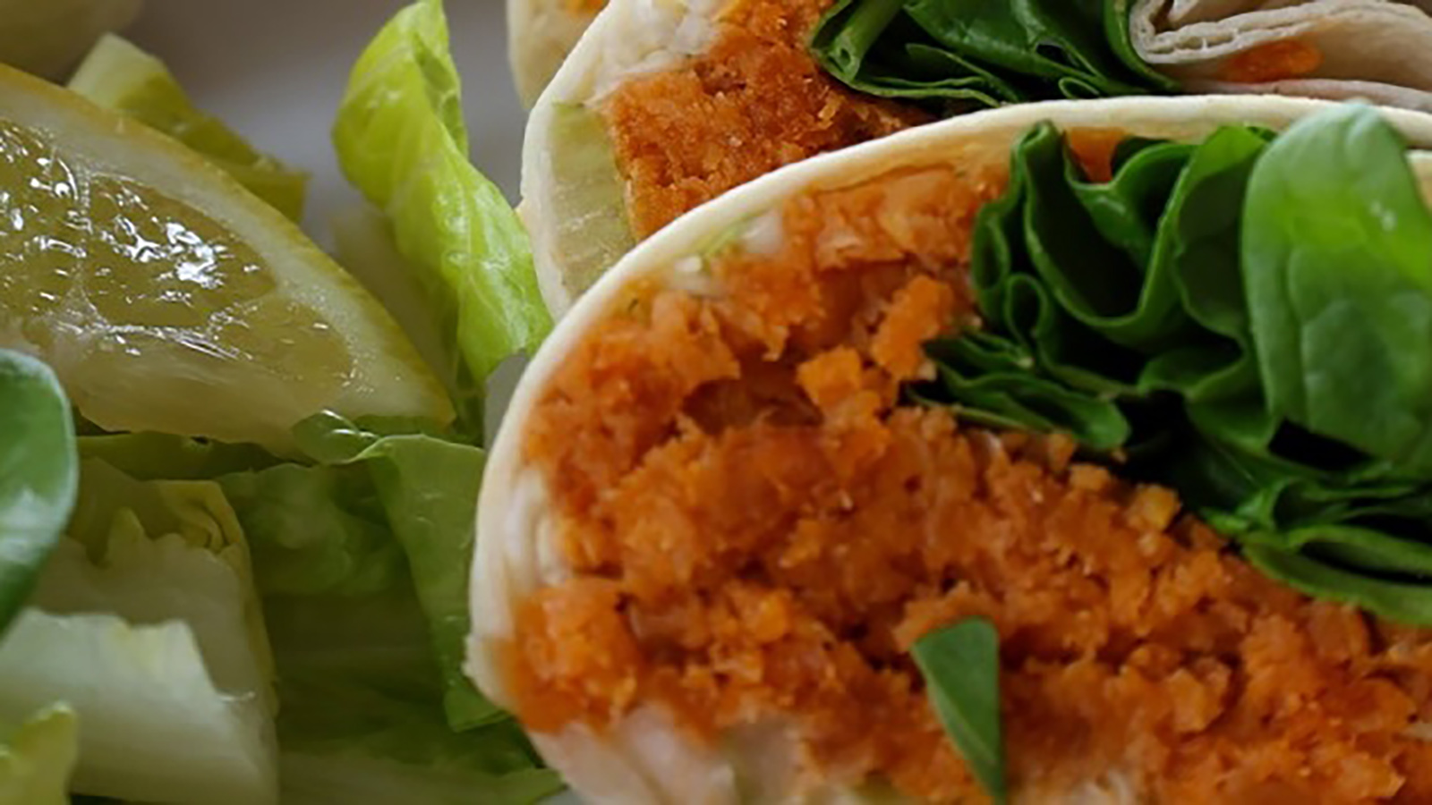 A close-up view of a wrap cut in half, showing a bright orange filling, possibly shredded or finely chopped, along with green leafy vegetables. The wrap is accompanied by slices of cucumber and pickled vegetables.