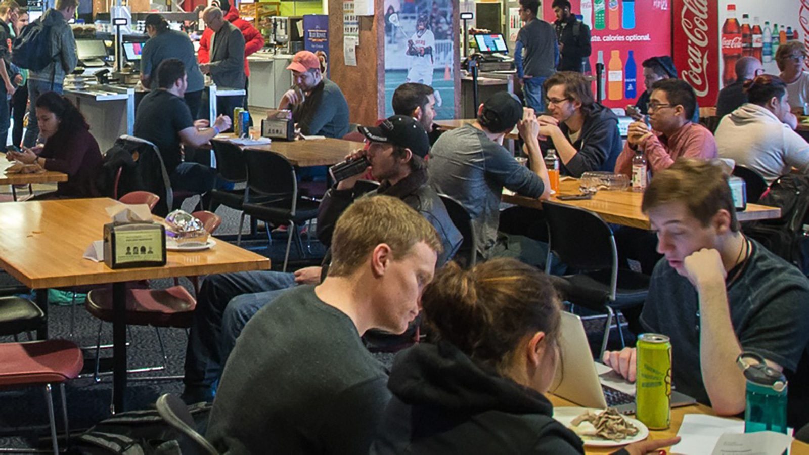 A busy cafeteria or dining area with multiple individuals seated at various tables. Some are engaged in conversation, while others are focused on laptops or personal devices. Vending machines and promotional posters are visible in the background, suggesting a casual, public space likely associated with a campus or corporate environment.