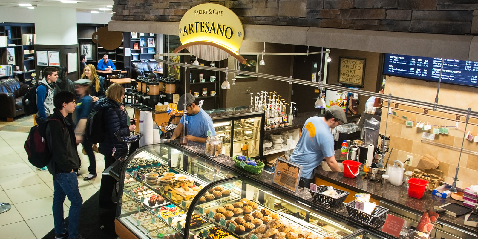 ﻿A bustling café, Artesano, features a vibrant display of pastries and beverages, with staff serving customers in a cozy, student-friendly atmosphere