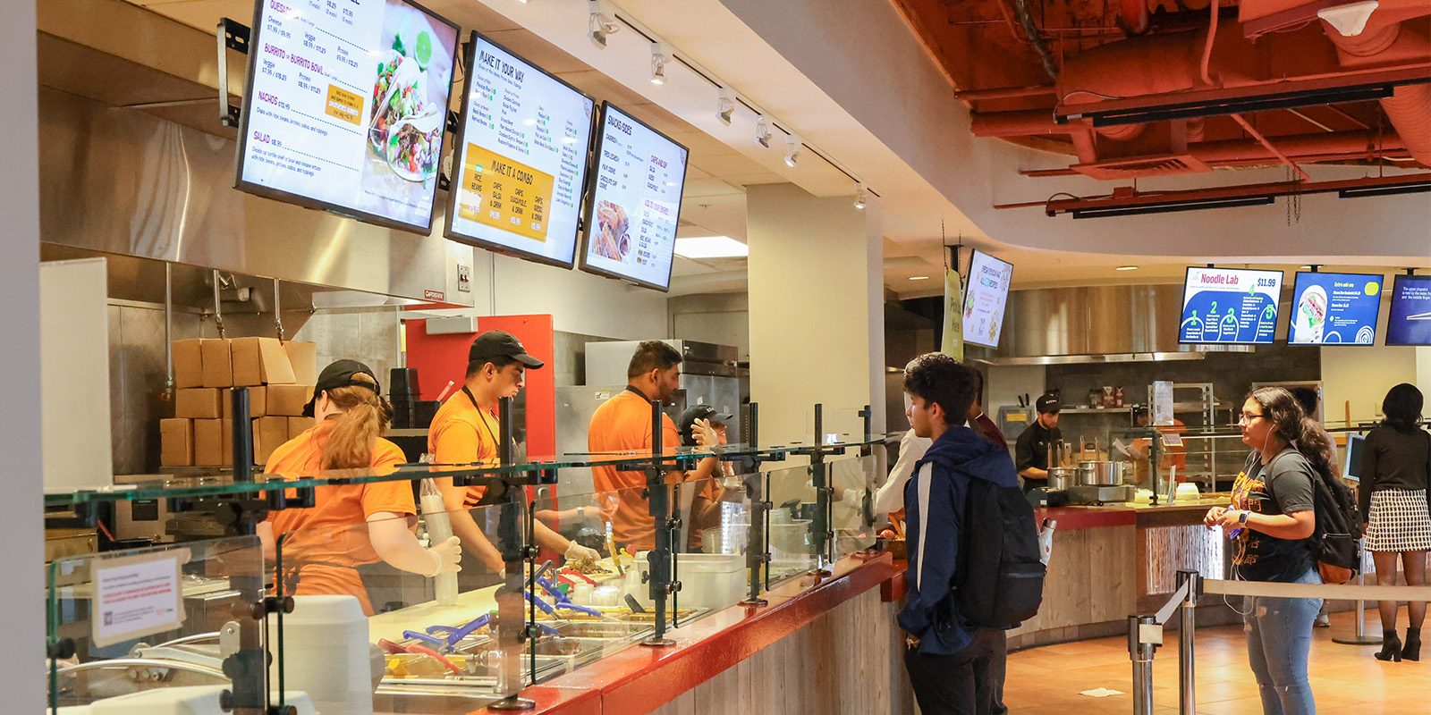 ﻿Food ordering station with employees making the food and customers in line inside the dining hall