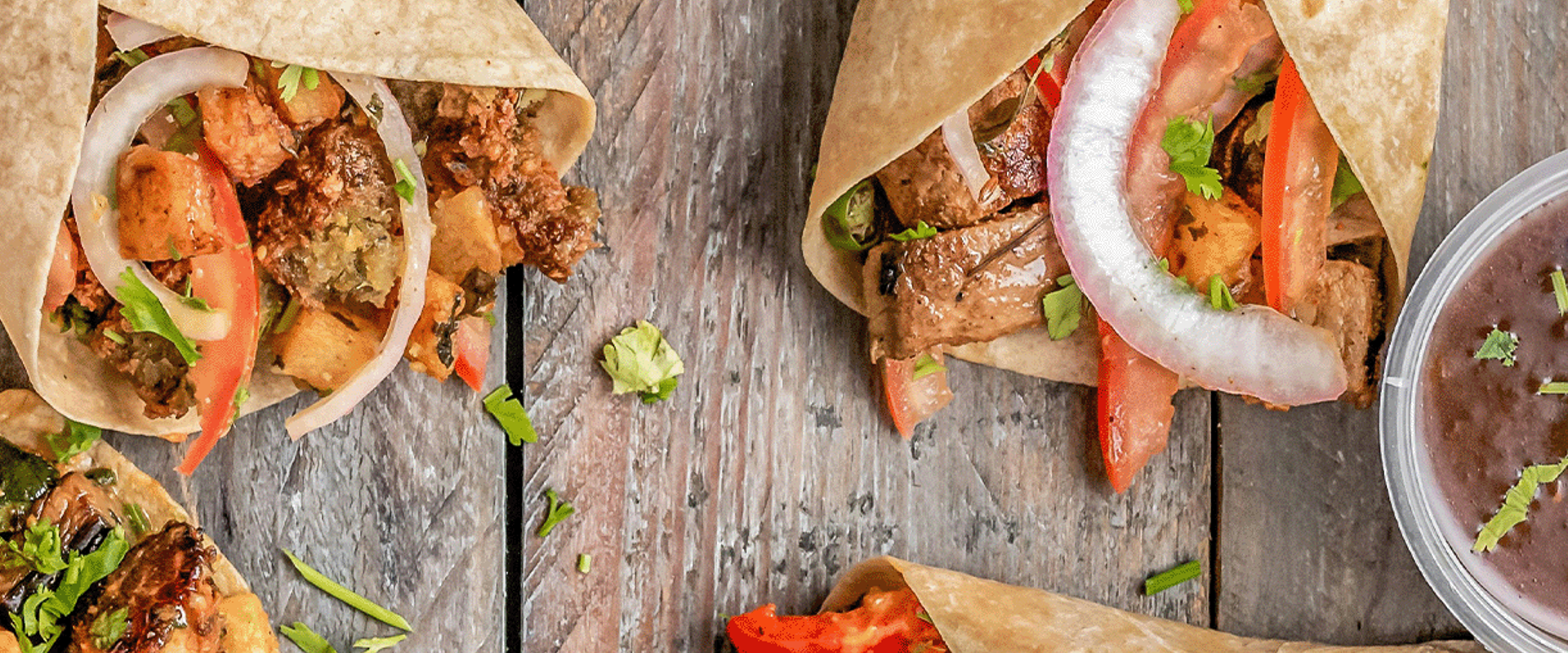 A close-up of a taco with slices of meat, tomato, onion, and cilantro wrapped in a soft tortilla. Next to the taco is a small container of black bean soup garnished with cilantro.