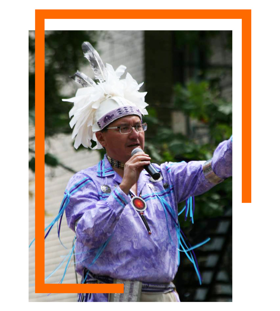 man in traditional Native outfit speaking into microphone with orange bar around photo