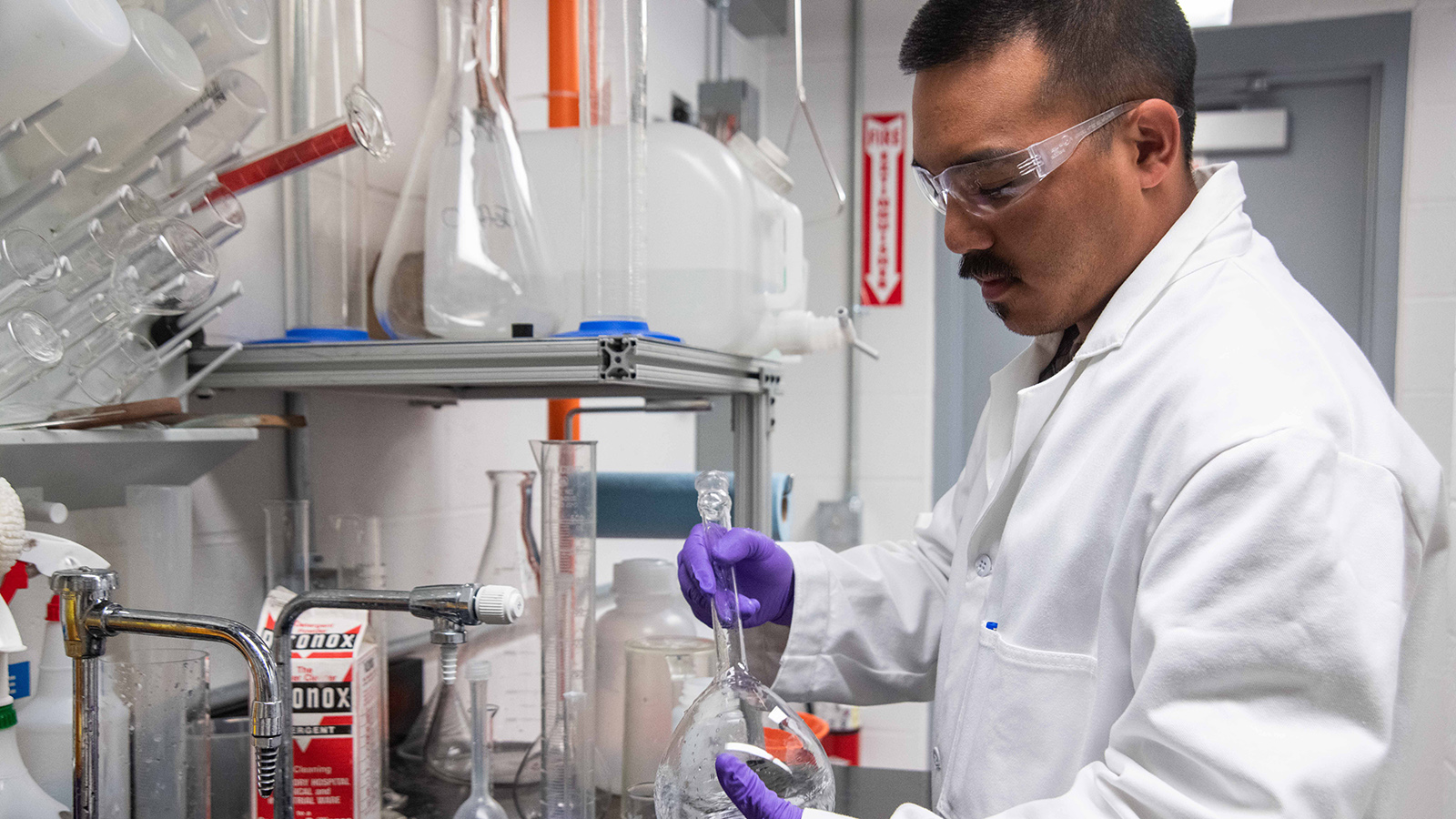 An individual in a white lab coat using a pipette to transfer a purple substance in a laboratory setting, surrounded by various glassware and containers.