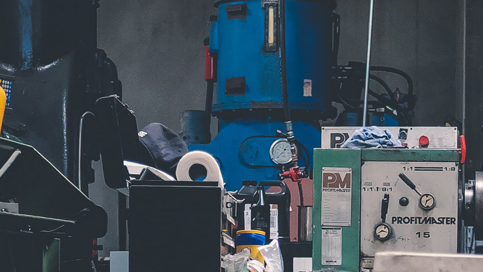 Industrial workshop with various machinery, including a large blue machine with gauges and valves, and a piece of equipment labeled “PROFIMASTER 1.5”.