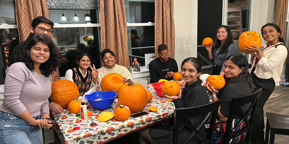 Nine people carving pumpkins around a dining table decorated with a fall-themed tablecloth, engaging in a festive pumpkin carving activity.