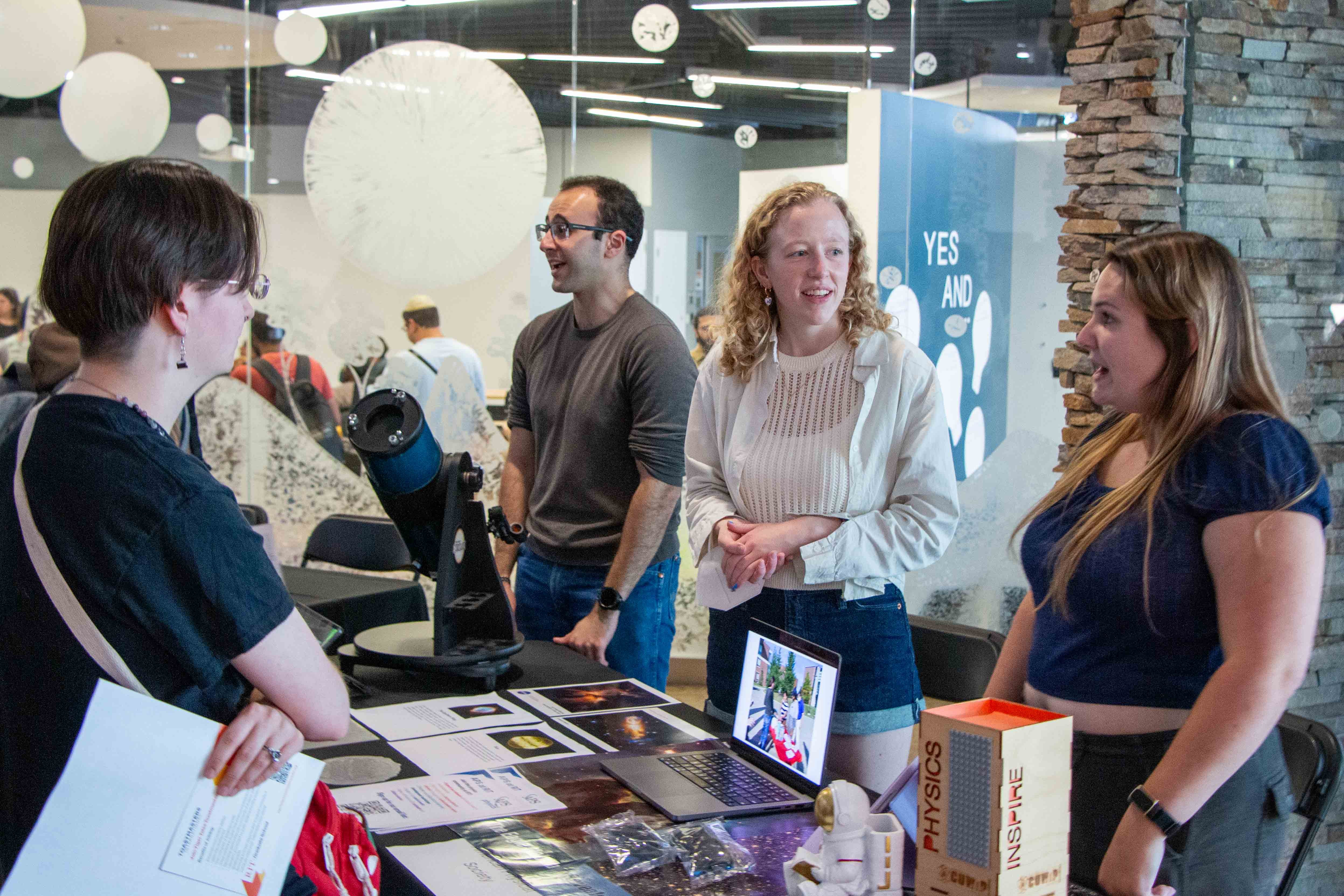 New graduate students sitting on a circular table with one student holding up and showing a gray RIT T-shirt while others smile at the camera