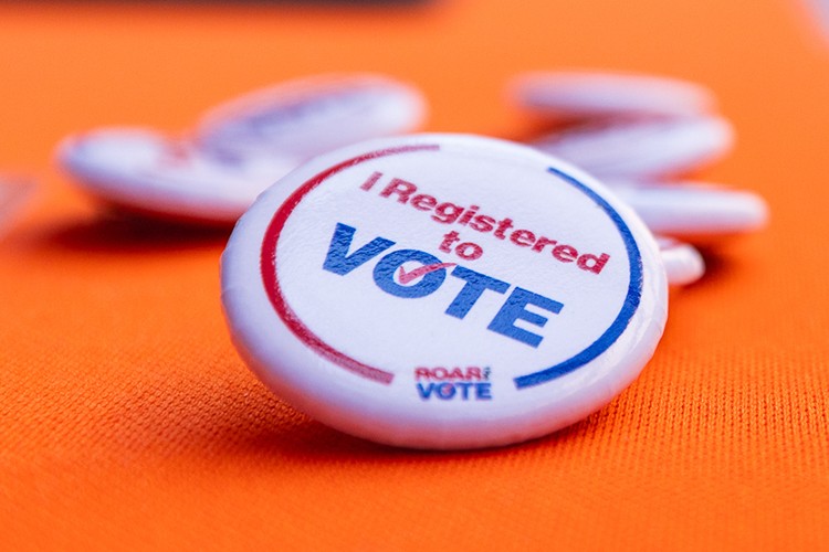A white button with red and blue text appears on the top of table with an orange table cloth covering it. The text on the button reads, "I registered to vote" with the Roar the Vote logo.