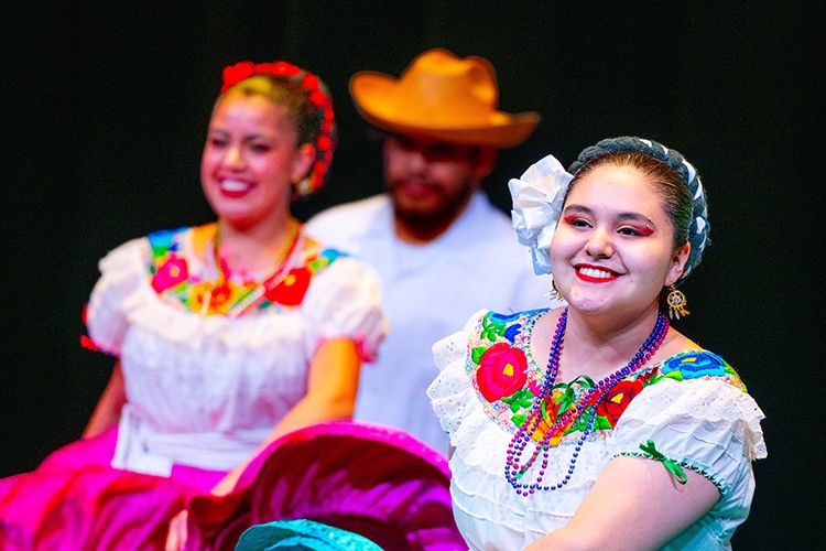 Students dressed in bright colored cultural apparel are shown dancing on stage