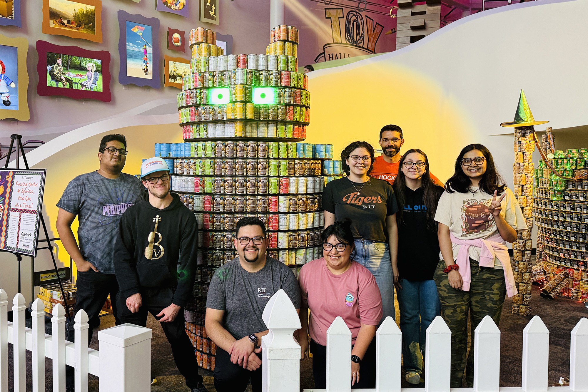 Group of eight people standing in front of a large, playful sculpture made from canned food, with each person smiling and wearing casual clothing.