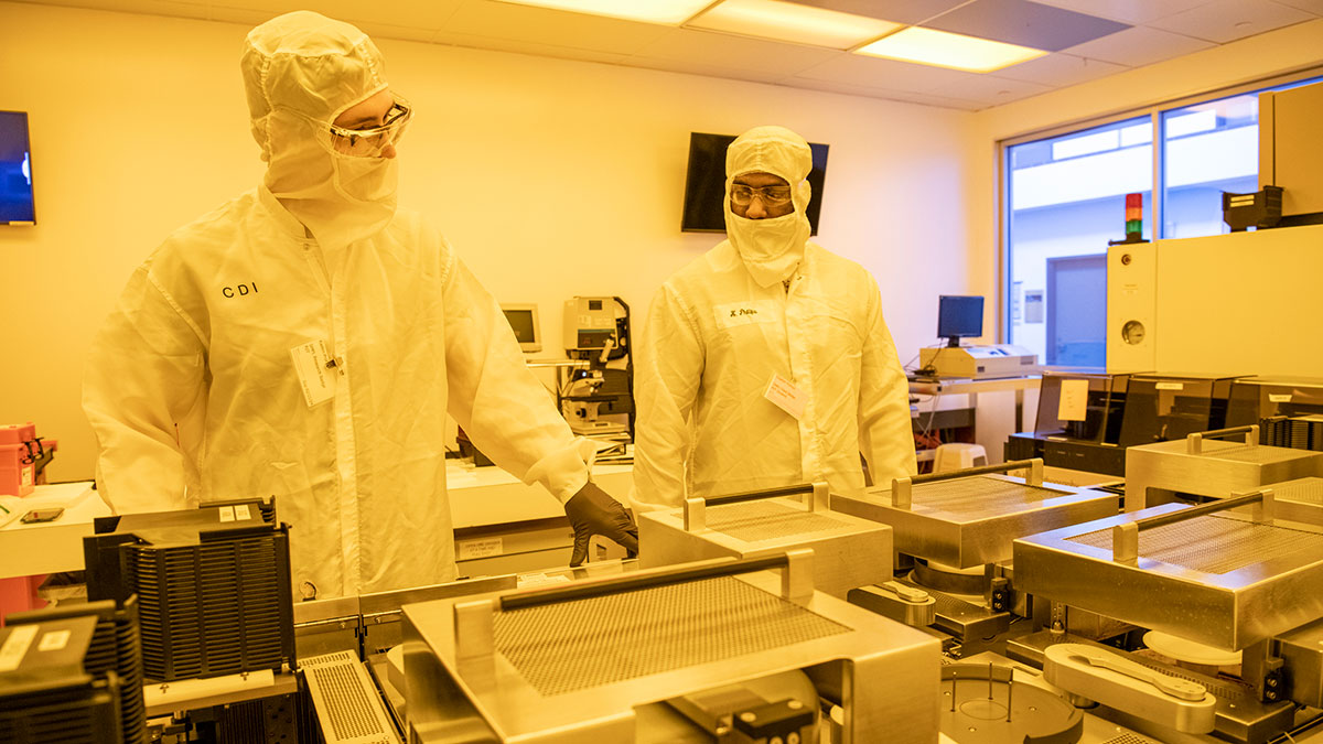 Two researchers in cleanroom suits work with semiconductor processing equipment in a high-tech lab.