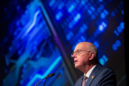 R I T President David Munson is shown in side profile from the chest up standing at a podium. Behind him, a screen is lit up with various shares of blue resembling a computer chip.