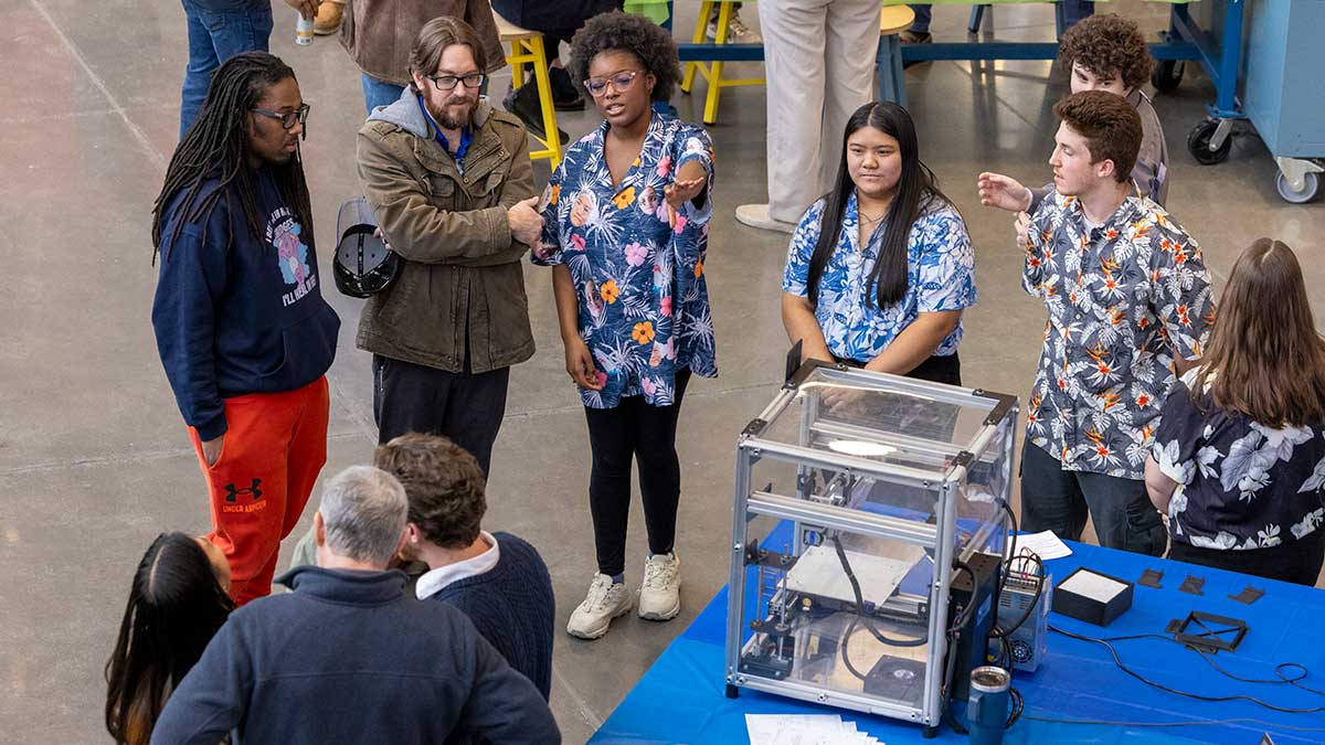 A group of students and mentors discussing a STEM project at an engineering exhibition, with a 3D printer on display.