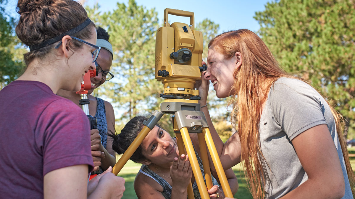 Three students outside looking into a surveyor scope.