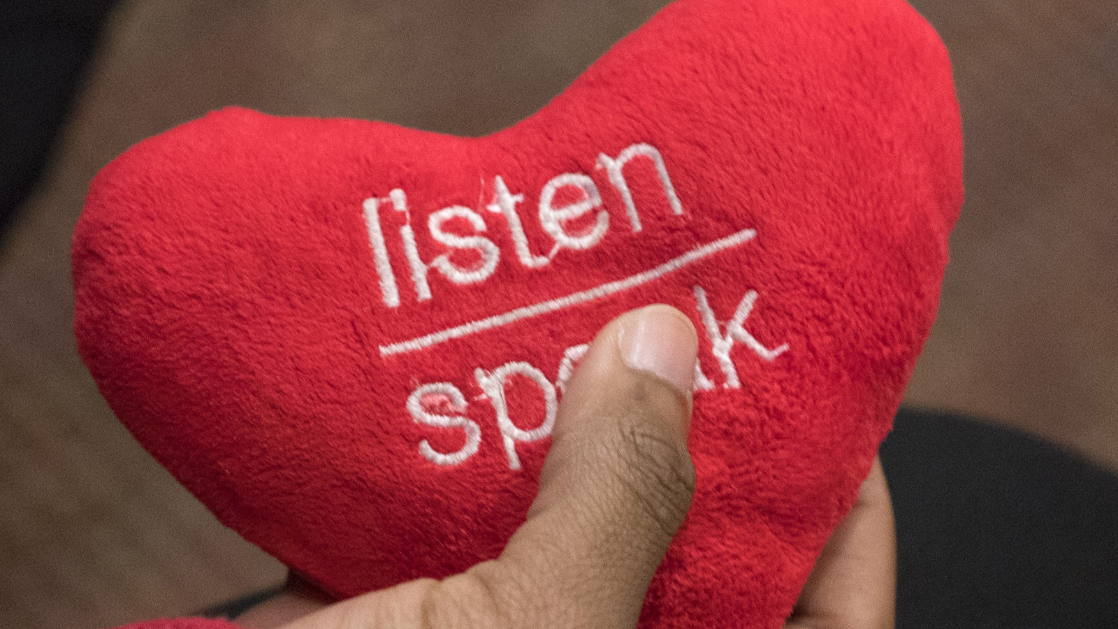 A hand holding a red heart-shaped object with the words “listen” and “spark” embroidered in white thread.