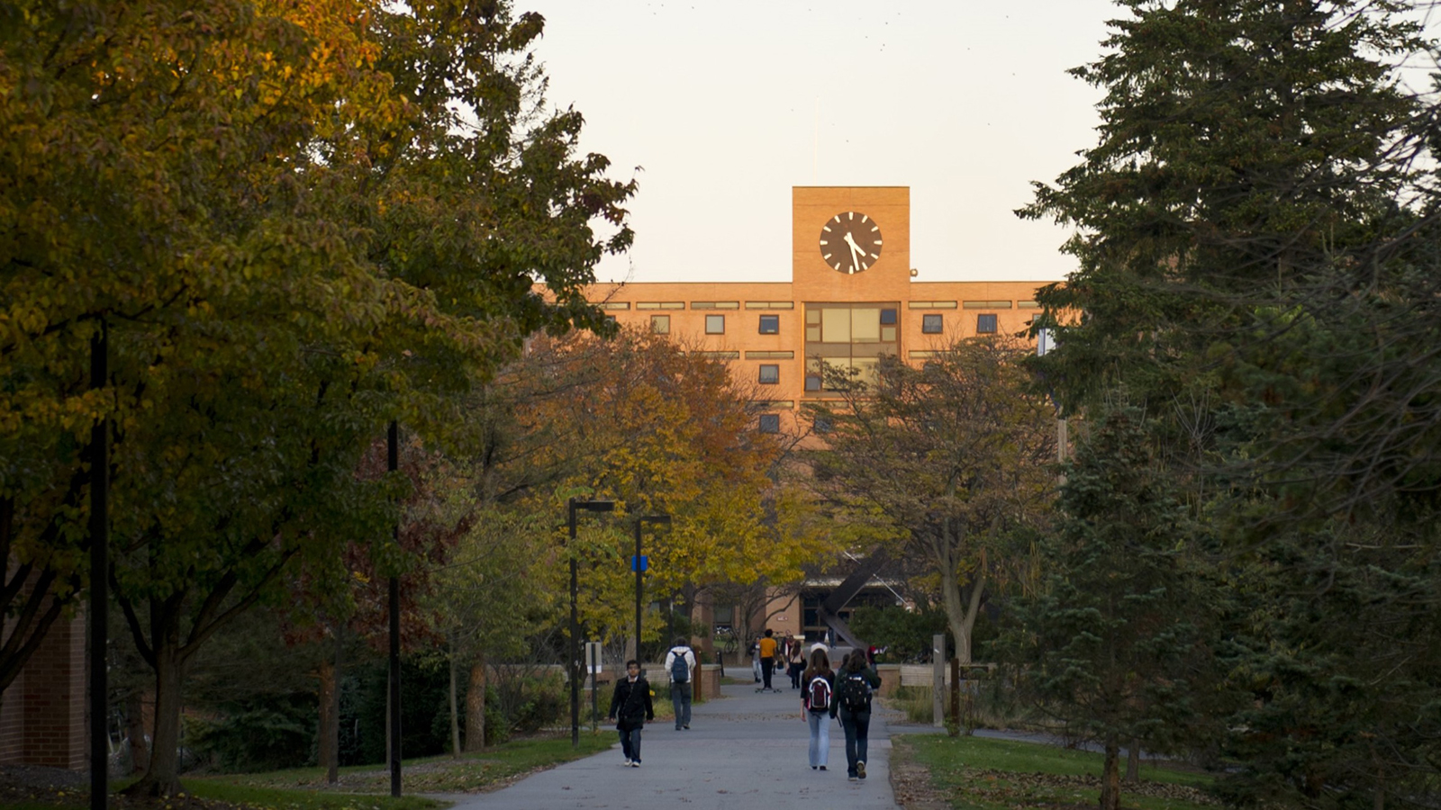 RIT campus during autumn with students walking on a pathway, trees with fall foliage, and a building with a large clock in the background.