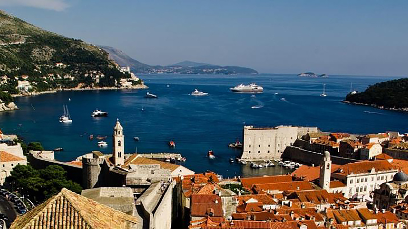 A panoramic view of Dubrovnik, Croatia, showcasing the historic old town with terracotta rooftops, the Adriatic Sea with various boats and yachts, and distant hills under a clear blue sky.