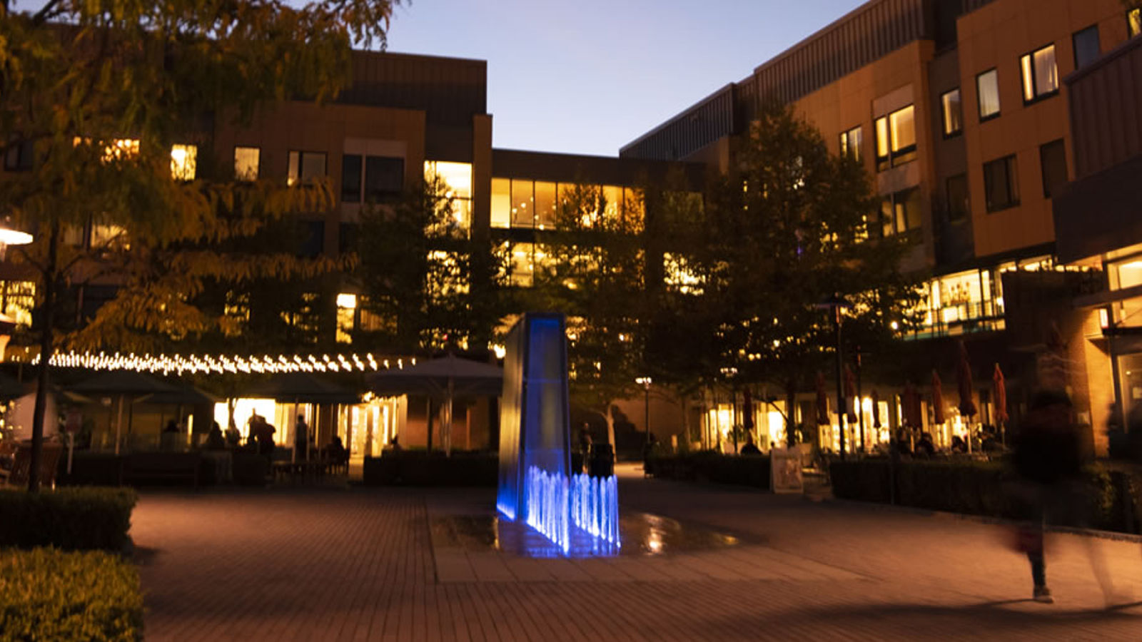 RIT's Global Village at dusk with a modern building featuring illuminated windows in the background, a paved plaza with benches and trees in the foreground, and a blue illuminated vertical water sculpture emitting light.