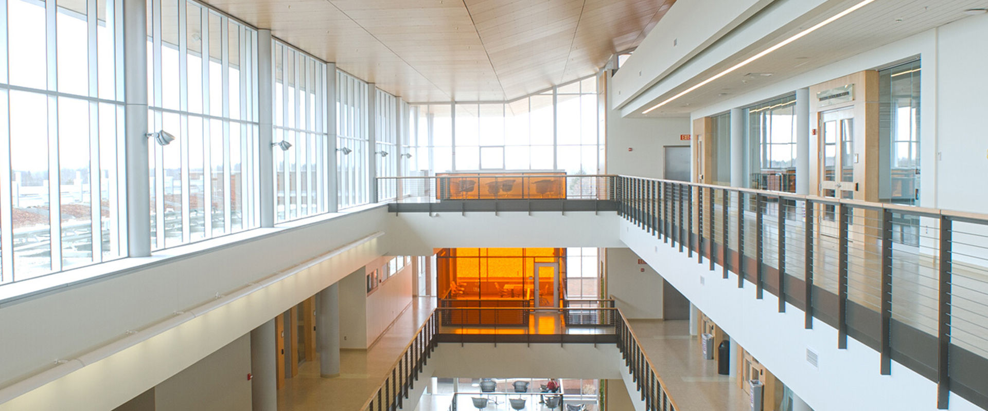 An interior view of a modern building with a spacious atrium, featuring large windows, a balcony on the upper level, and a distinctive orange glass enclosure.