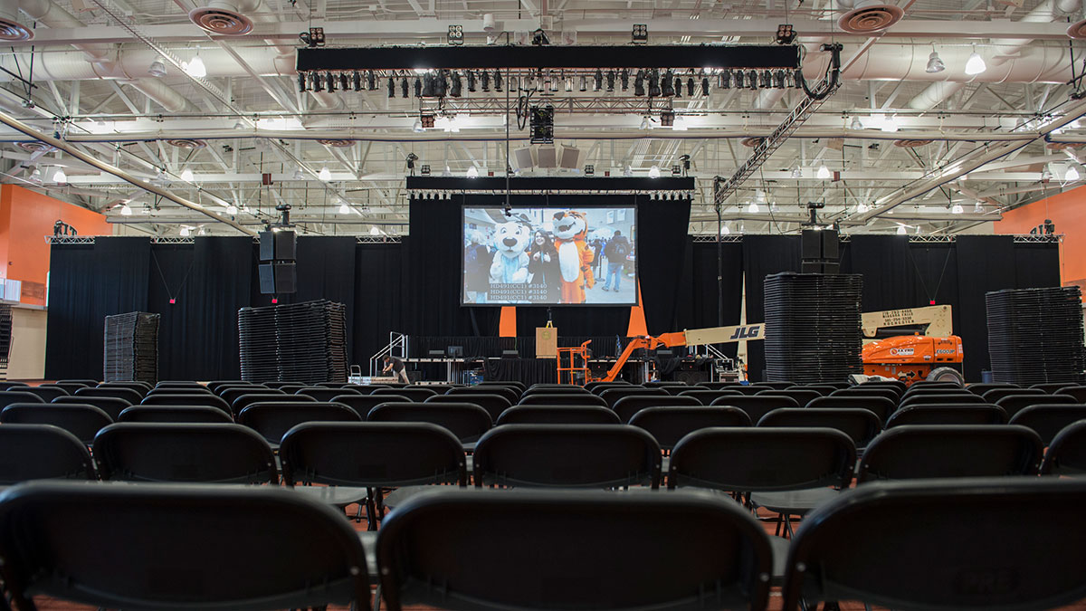 An empty event venue with rows of chairs, a stage, and a large screen displaying mascots on the screen.