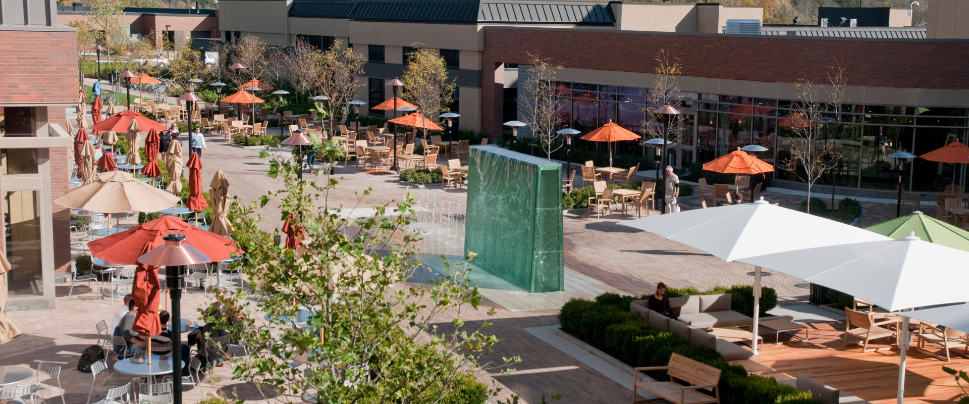 An outdoor seating area with wooden chairs and tables, green umbrellas, and people walking in the background near RIT's Salsarita's restaurant in Global Village.